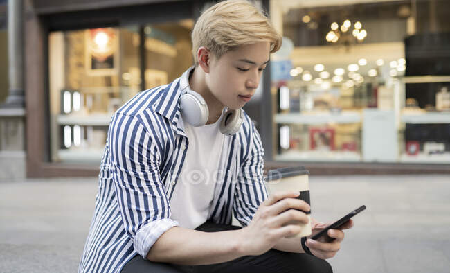 Young ethnic male with blond hair browsing mobile phone while sitting in street with takeaway beverage — Stock Photo