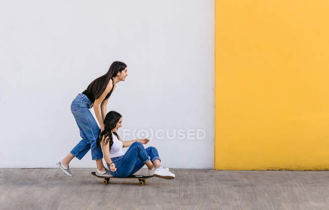 Cheerful teen with crossed legs riding skateboard with content female sibling on walkway in daytime — Stock Photo