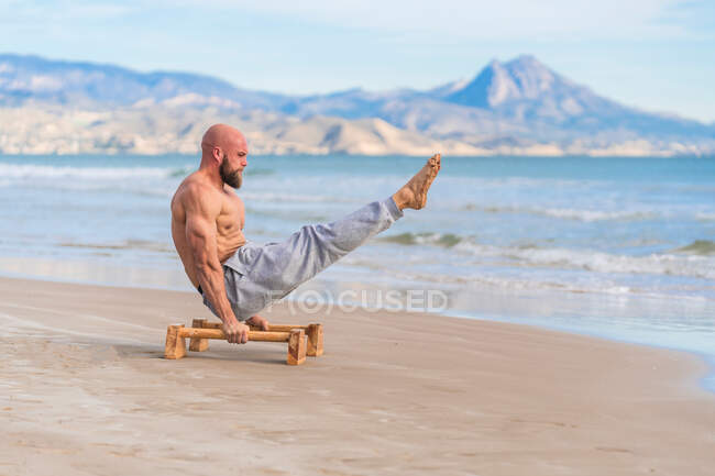 Side view of focused bald strong athlete working out lifting up his own wait from bars on sandy coast — Stock Photo
