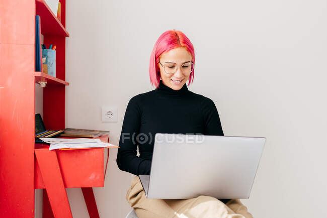 Freelancer jovem focado em roupas casuais sentadas na cadeira e usando laptop enquanto trabalhava no projeto em apartamento moderno leve — Fotografia de Stock