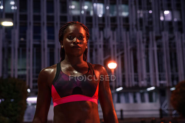 Ethnique musclé jolie femme en tenue de sport debout sur la rue dans le crépuscule et regardant la caméra — Photo de stock
