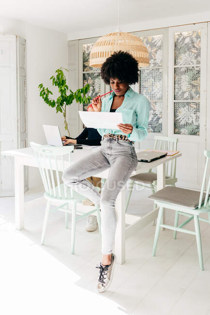 Moderno bem sucedido Africano americano feminino freelancer em roupa elegante com cabelo afro sentado à mesa e documento de leitura em casa — Fotografia de Stock