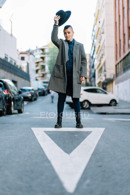 Transgender person in stylish coat and hat standing with raised arm on urban roadway while looking at camera — Stock Photo