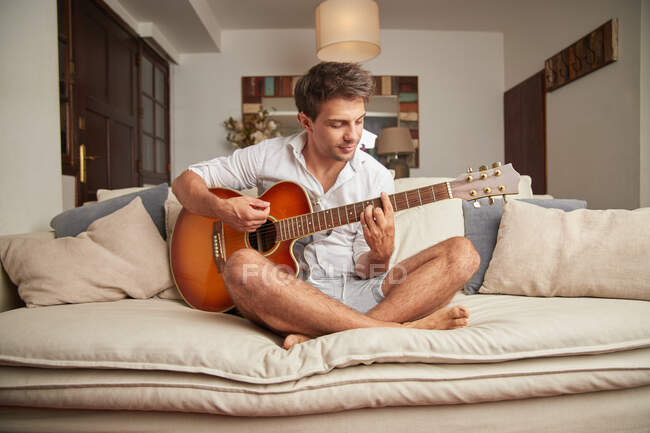 Дорослий чоловік у повсякденному одязі, сидячи на дивані, грає на акустичній гітарі у світлій вітальні — стокове фото