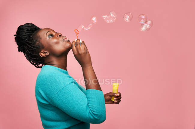 Seitenansicht einer fröhlichen Afroamerikanerin, die in blauer Kleidung wegschaut und im Studio Seifenblasen vor rosa Hintergrund pustet — Stockfoto