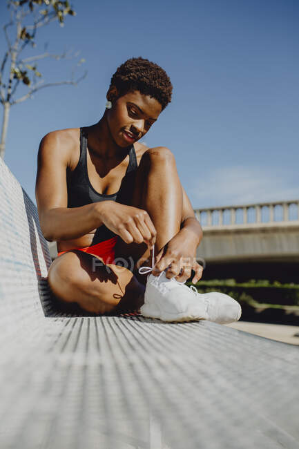 Young fitness African American woman resting and tying the laces after making exercise on park bench — Stock Photo