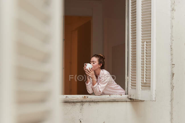 Tranquilo fêmea no pijama inclinado na janela com xícara de café da manhã e olhos fechados — Fotografia de Stock
