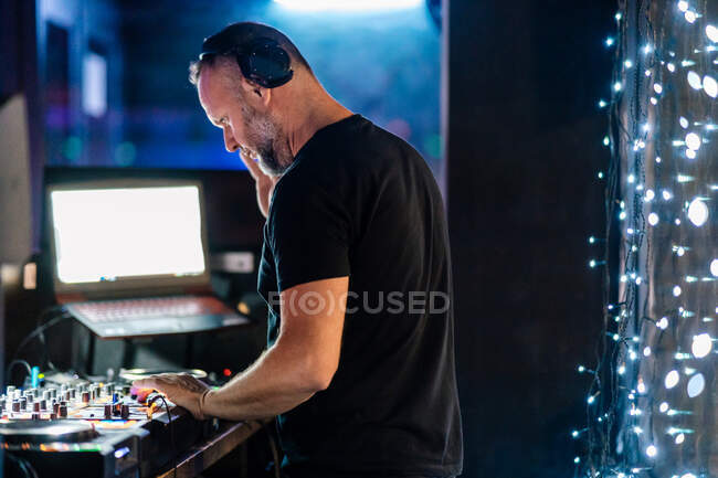 Side view pensive middle aged man in headphones and black casual clothes focusing and playing professional DJ controller while performing at concert in contemporary night club against laptop with blank screen — Stock Photo