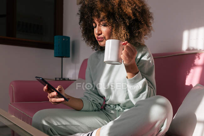 Афроамериканка просматривает мобильный телефон и пьет горячий кофе, отдыхая на диване в выходные дни дома — стоковое фото