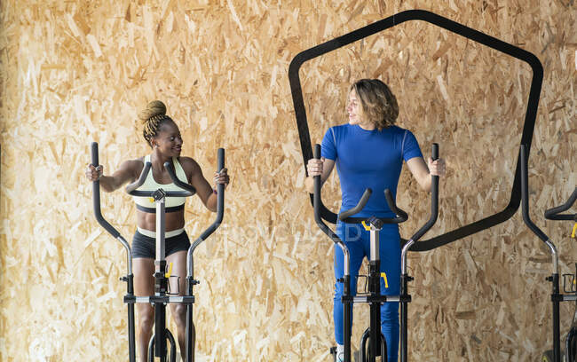 Atletas multirraciales sonrientes con ropa deportiva haciendo ejercicio en bicicletas estáticas mientras hablan y se miran en el gimnasio - foto de stock