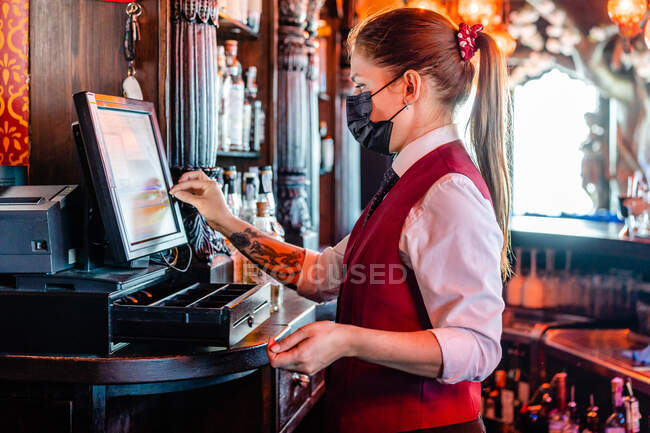 Vue latérale du barman femelle dans un masque de protection à l'aide d'une caisse et d'un écran tactile pendant qu'elle travaille dans une barre pendant le coronavirus — Photo de stock
