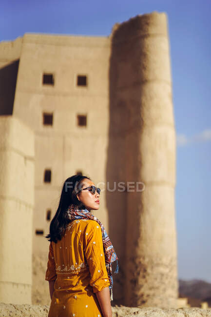 Обратный вид молодой азиатской путешественницы в красочной одежде и солнцезащитных очках, стоящих против средневековья: замок Бахла и взгляд в сторону во время отпуска в Омане — стоковое фото