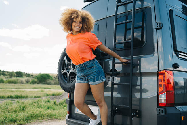 Mujer afroamericana optimista con el pelo rizado sonriendo y mirando hacia otro lado mientras agarra la escalera en la parte posterior de la casa rodante en el día de verano en el campo - foto de stock