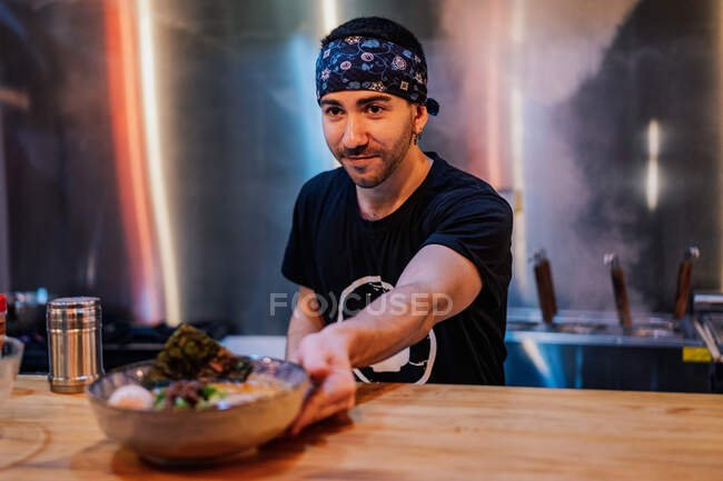 Bärtiger Mann mit Kopftuch und schwarzem T-Shirt legt Teller mit Nudeln auf Holztheke für Kunden in Ramen-Bar — Stockfoto