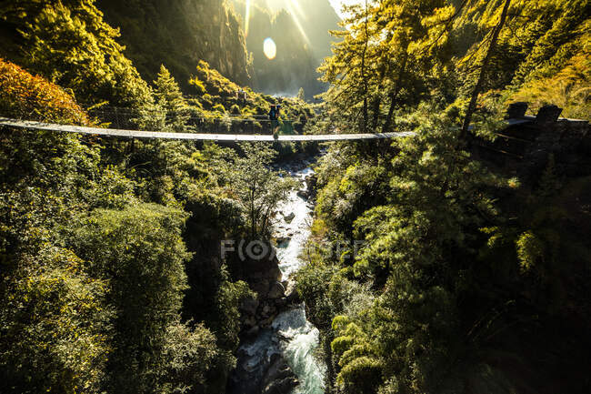 Explorador irreconhecível caminhando ao longo da ponte pedonal de suspensão metálica sobre o rio rápido na cordilheira do Himalaia, no Nepal — Fotografia de Stock