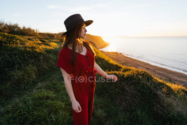 Attraktive junge Frau in rotem Kleid und Hut steht auf einer grünen Wiese in sonniger Landschaft — Stockfoto