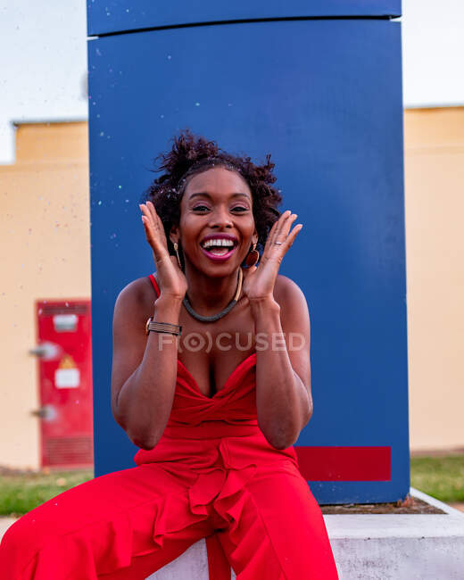 Joven mujer afroamericana alegre en traje rojo con peinado afro mirando a la cámara durante el día - foto de stock
