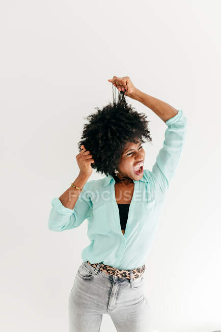 Verspielte junge Afroamerikanerin mit geschlossenen Augen im trendigen Outfit, die Spaß daran hat, Afrohaare auf weißem Hintergrund zu berühren — Stockfoto