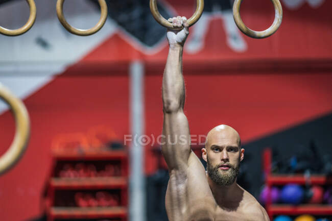 Сильний без сорочки чоловік дивиться на камеру, що стоїть, робить вправи на гімнастичних кільцях під час інтенсивного тренування в сучасному тренажерному залі — стокове фото