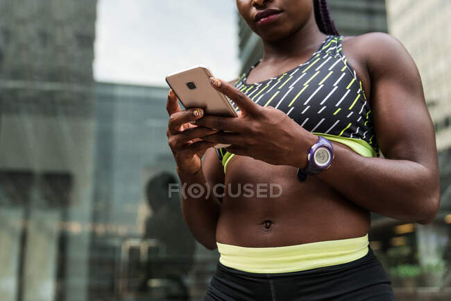 Ernte Afroamerikanerin in Sportbekleidung surft auf modernem Smartphone, während sie während des Trainings im Freien auf verschwommenem Hintergrund der Stadtstraße steht — Stockfoto