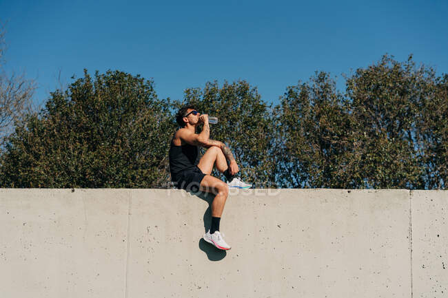 Вид сбоку спортсмена в спортивной одежде, пьющего аква из бутылки после тренировки против деревьев на солнце — стоковое фото