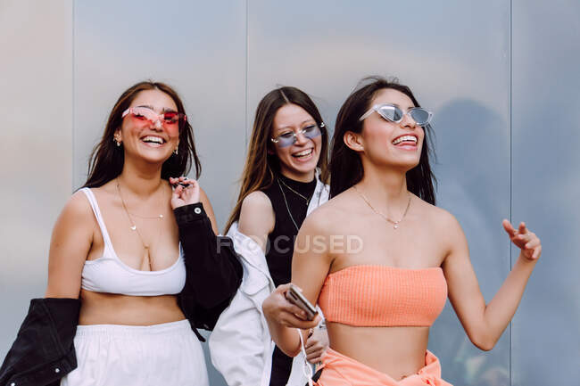Amigas positivas en gafas de sol de moda y ropa casual riéndose felizmente mientras pasan un rato agradable juntas - foto de stock