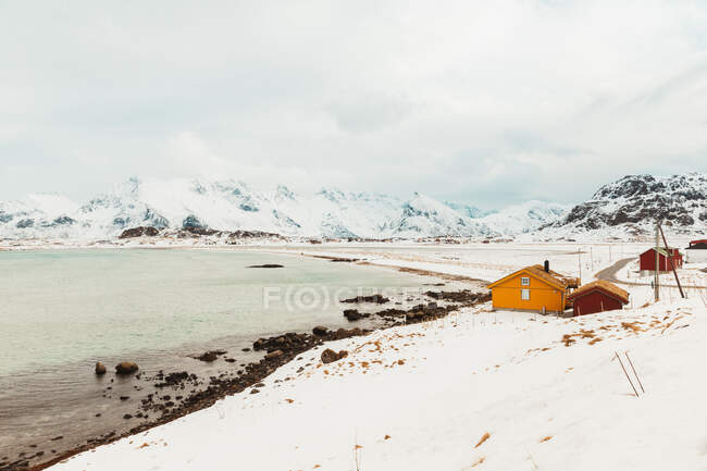 Wooden colorful shacks located on white snowy coast surrounded by mountain range on Lofoten Islands, Norway — Stock Photo