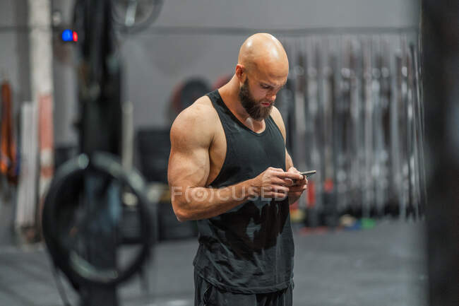 Vista lateral do desportista careca muscular em pé no ginásio moderno navegando no smartphone durante a pausa para treino — Fotografia de Stock