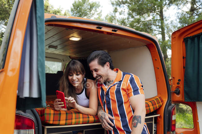 Захоплена пара мандрівників, сидячи у фургоні і знімаючи себе на смартфоні, обіймаючи і насолоджуючись літніми пригодами — стокове фото