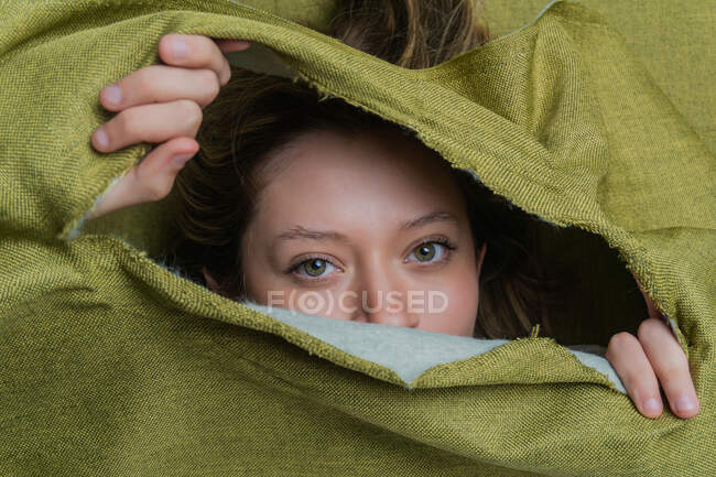 Jeune femelle aux yeux verts regardant la caméra tout en se cachant derrière un tissu vert déchiré — Photo de stock