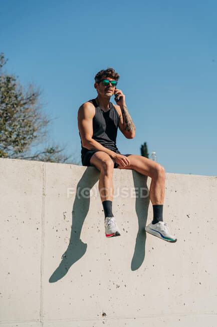 Подходящий спортсмен в спортивной одежде разговаривает по мобильному телефону, отдыхая на заборе после тренировки — стоковое фото