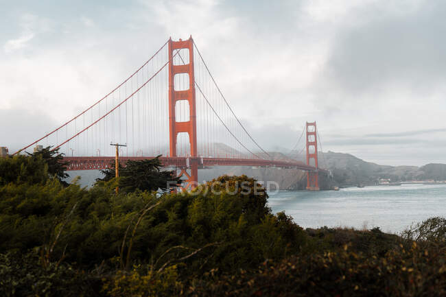 Ponte Golden Gate famosa pendurada acima do rio com a costa verde sob o céu cinzento sombrio em San Francisco — Fotografia de Stock