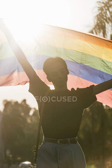 Deleitado macho gay de pie con los ojos cerrados levantando arco iris LGBT bandera durante el atardecer en la ciudad - foto de stock