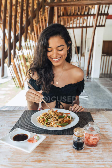 Cheerful female tourist with delicious pasta between food sticks above table with soy sauce and pickled ginger slices outdoors — Stock Photo