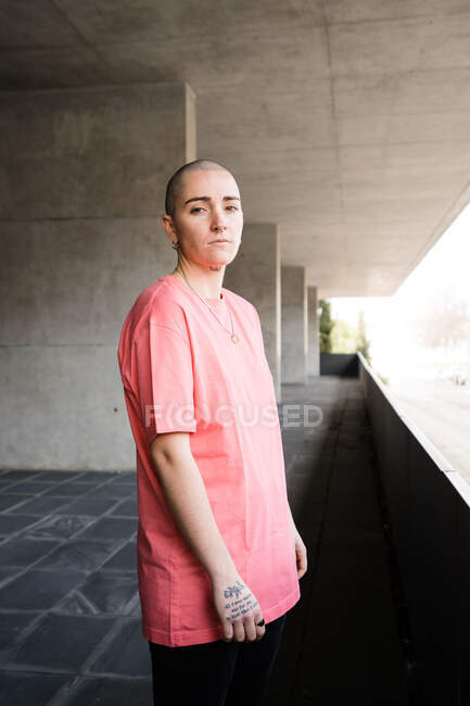 Side view of transgender person in t shirt standing looking at camera against fence in masonry construction in daytime — Stock Photo