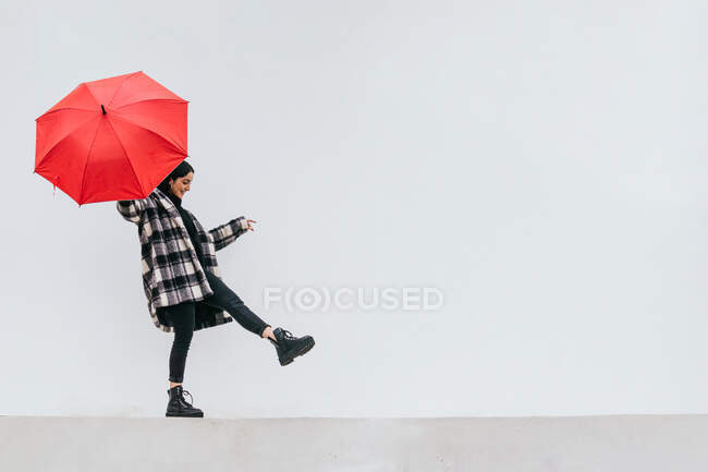 Fröhliches junges Weibchen mit rotem Regenschirm läuft und balanciert an einem regnerischen Tag auf der Straße gegen eine graue Wand — Stockfoto