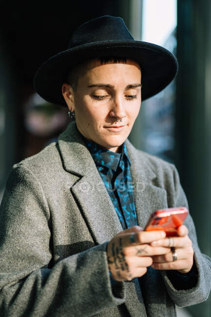 Androgynous pessoa em chapéu navegando no celular olhando para a tela de pé na rua à luz do dia — Fotografia de Stock