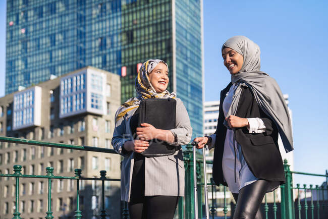 Des femmes entrepreneurs musulmanes souriantes avec une valise marchant le long de la rue en ville et se regardant — Photo de stock
