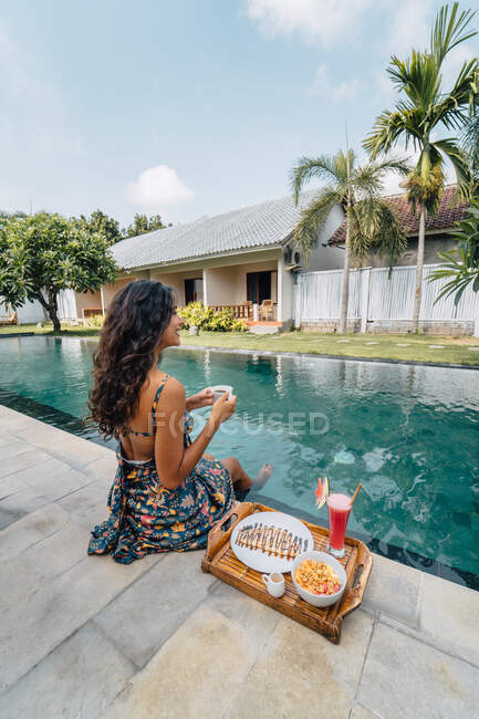 Вид ззаду весела жінка-мандрівник з гарячим напоєм, дивлячись далеко, сидячи на березі басейну зі смачним сніданком на підносі — стокове фото