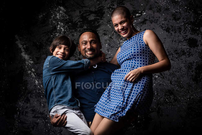 Encantado homem étnico segurando mulher e filho e se divertindo no estúdio no fundo escuro — Fotografia de Stock
