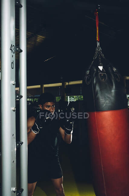Joven centrado hombre asiático entrenamiento boxeo realizar golpes mientras se ejercita con saco de boxeo pesado en un gimnasio moderno - foto de stock