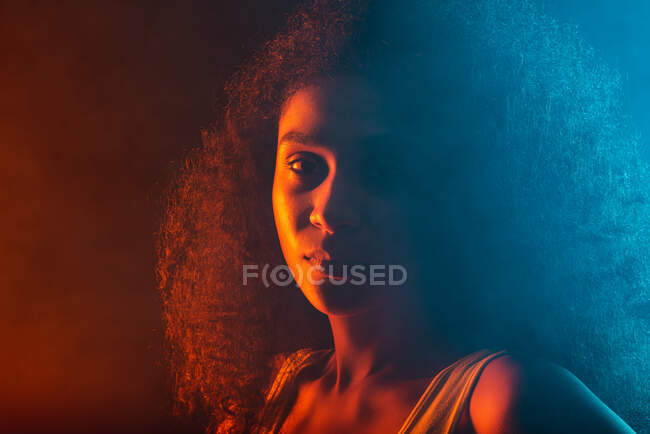 Junge Afroamerikanerin mit lockigem Haar blickt in die Kamera auf schwarzem Hintergrund mit Neonlicht — Stockfoto