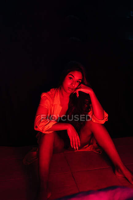 Чувственная молодая латиноамериканка в нижнем белье смотрит в камеру, отдыхая на террасе под красным неоном — стоковое фото