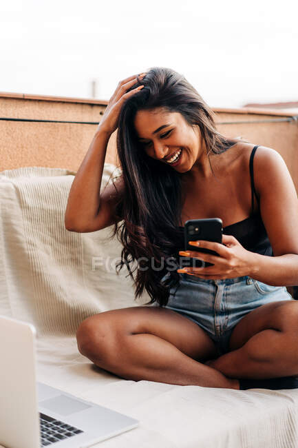 Jovem hispânica otimista tocando o cabelo e rindo enquanto se senta no sofá perto do laptop e do smartphone de navegação na varanda — Fotografia de Stock