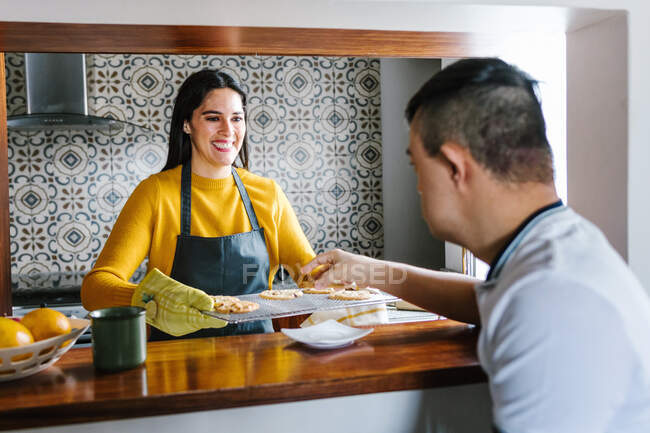 Cheerful ethnic mother giving hot baked cookies for teenage boy with Down syndrome in kitchen at home — Stock Photo