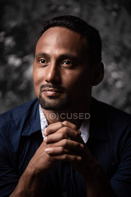 Delighted stylish ethnic male leaning on wooden stool in studio on dark background and looking away — Stock Photo