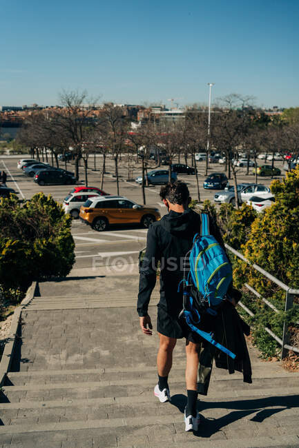 Back view of anonymous male athlete in sports clothes with rucksack strolling on city stairs against parking — Stock Photo
