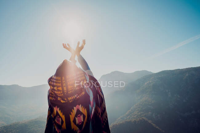 Обратный вид на неузнаваемую женщину в традиционной одежде, занимающуюся йогой в горной позе с поднятыми руками, стоя в горах в солнечный день — стоковое фото