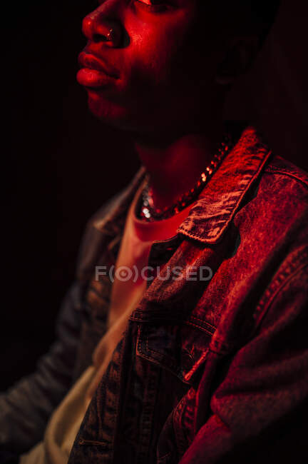 Crop calmo homem afro-americano elegante em jaqueta de jeans sob luz vermelha néon na sombra no fundo preto olhando para longe — Fotografia de Stock