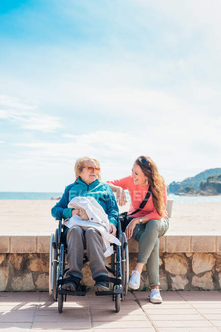 Fröhliche erwachsene Tochter mit älterer Mutter im Rollstuhl sitzt im Sommer am Steinzaun entlang der Strandpromenade — Stockfoto
